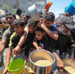 Crowd of Palestinian men and boys in Gaza urgently reaching for large pots of food during a community distribution of meals, with many holding empty buckets and bowls.