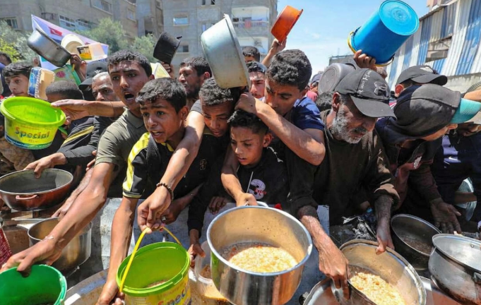 Crowd of Palestinian men and boys in Gaza urgently reaching for large pots of food during a community distribution of meals, with many holding empty buckets and bowls.